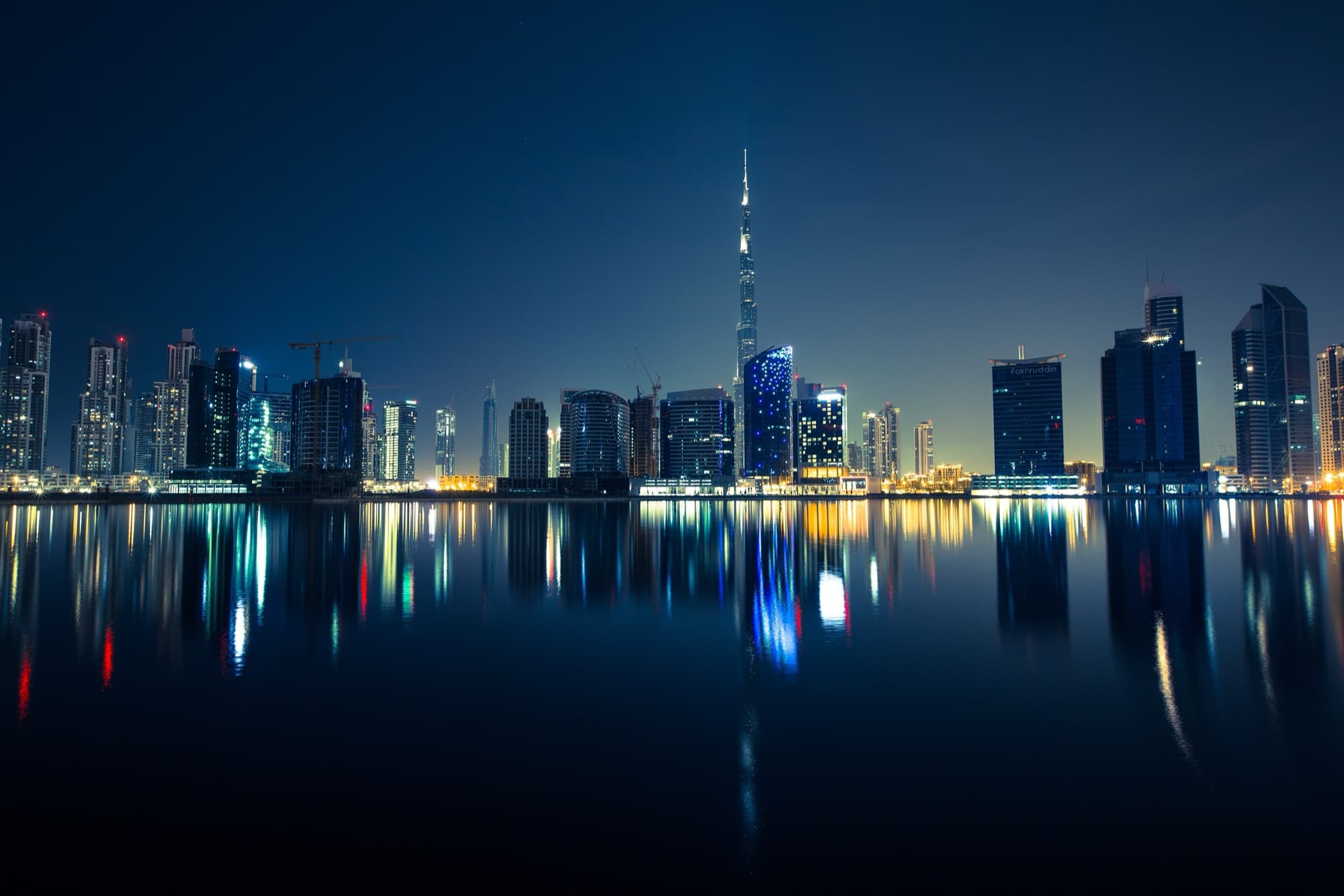 The Dubai skyline and Burj Khalifa lit up at night, reflected in calm water.
