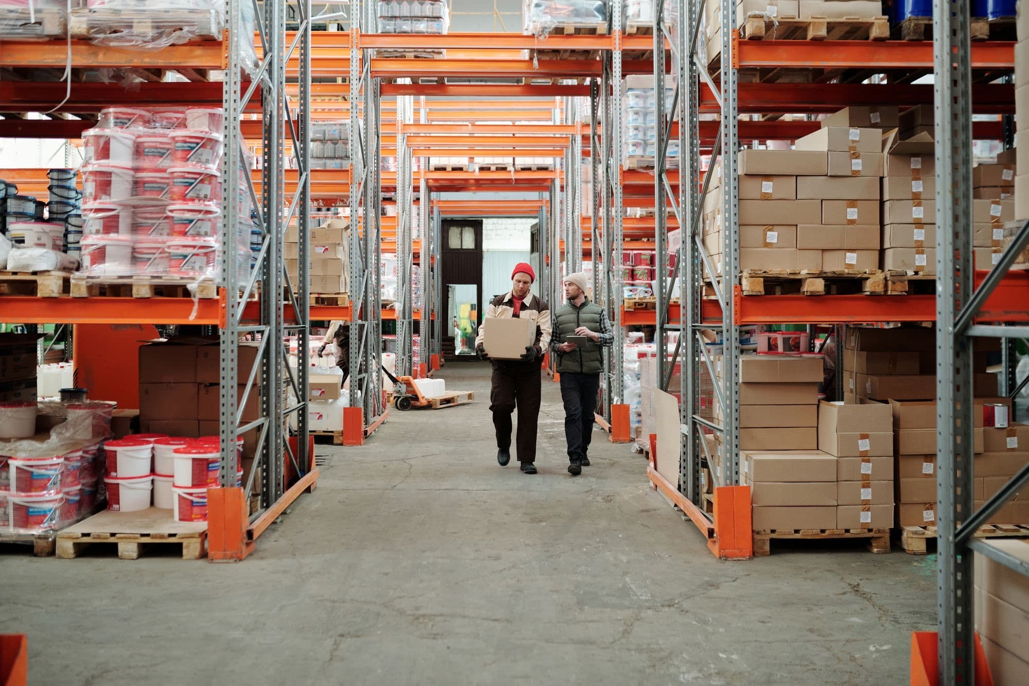 Two warehouse workers walk down a pallet-rack aisle carrying a carton for an order.