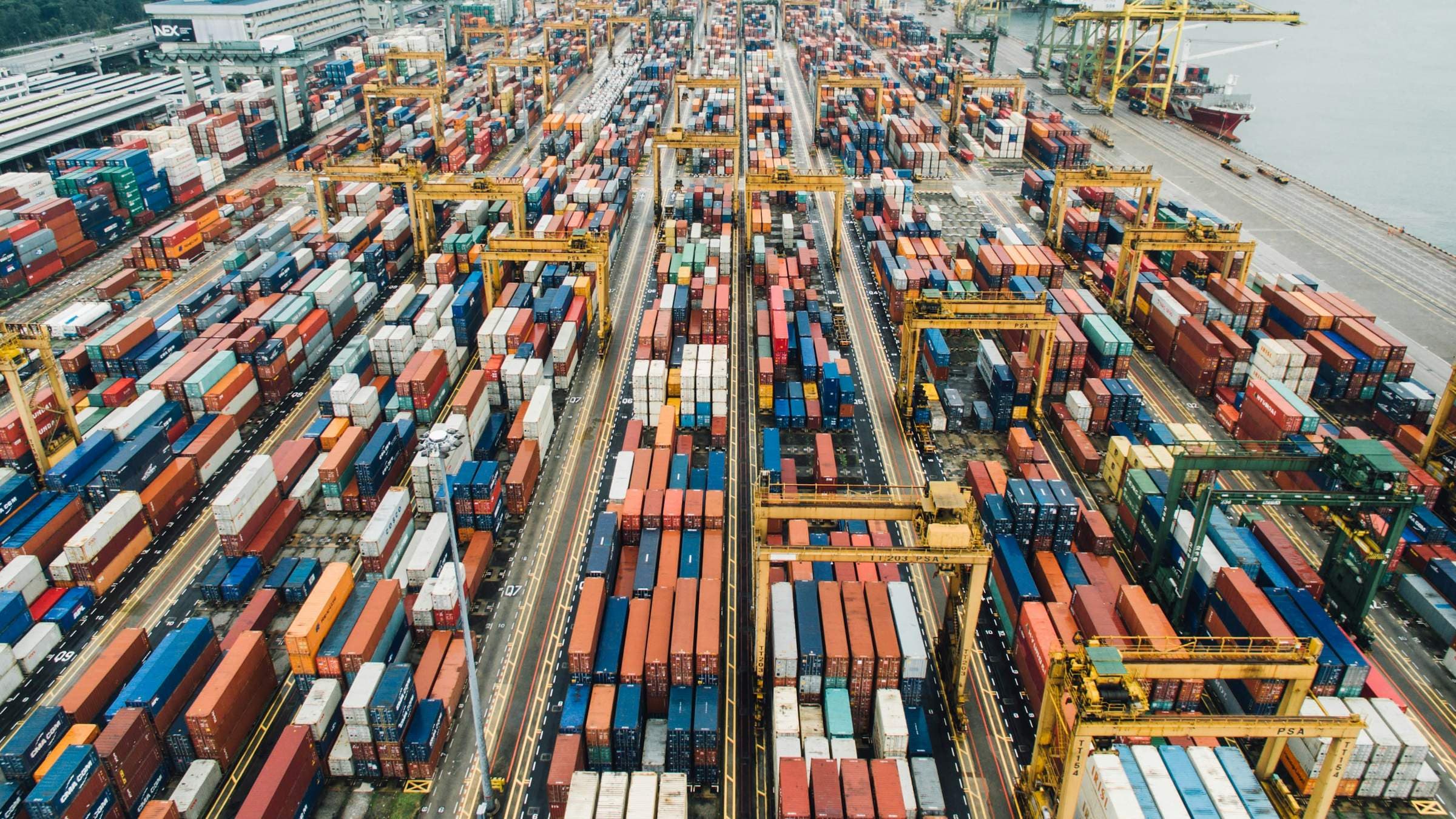 Stacked shipping containers on a container terminal quay, with gantry cranes loading a vessel.